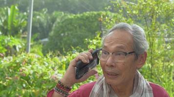 Elderly grandfather talking to his friend on mobile phone in the backyard at home.
