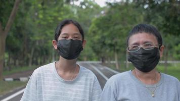 Mother and her daughter wears black face mask walking together on the footpath in public park.