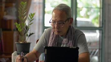hombre mayor escribiendo en un bloc de notas y trabajando en una tableta digital en casa.