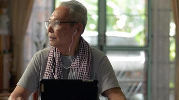 Senior man looking away to the window while listening social media online with earphones on digital tablet at home. video