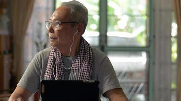 Senior man looking away to the window while listening social media online with earphones on digital tablet at home. video