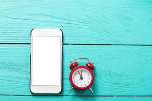 Mobile phone and red retro alarm clock with five minutes to twelve o'clock on blue wooden background. Top view and blank screen photo