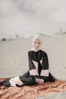 hermosa modelo femenina islámica usando moda hijab, un vestido de novia moderno para mujer musulmana sentada en la arena y la playa. retrata a una modelo asiática usando hiyab divirtiéndose en la playa con árboles foto