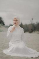 Beautiful islamic female model wearing hijab fashion, a modern wedding dress for muslim woman sitting in the sand and beach. Portrait an asian girl model using hijab having fun at the beach with trees photo