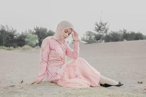 hermosa modelo femenina islámica usando moda hijab, un vestido de novia moderno para mujer musulmana sentada en la arena y la playa. retrata a una modelo asiática usando hiyab divirtiéndose en la playa con árboles foto
