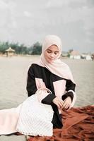 Beautiful islamic female model wearing hijab fashion, a modern wedding dress for muslim woman sitting in the sand and beach. Portrait an asian girl model using hijab having fun at the beach with trees photo