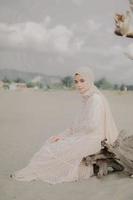 Beautiful islamic female model wearing hijab fashion, a modern wedding dress for muslim woman sitting in the sand and beach. Portrait an asian girl model using hijab having fun at the beach with trees photo