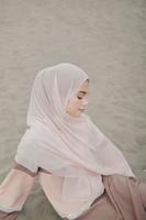 hermosa modelo femenina islámica usando moda hijab, un vestido de novia moderno para mujer musulmana sentada en la arena y la playa. retrata a una modelo asiática usando hiyab divirtiéndose en la playa con árboles foto