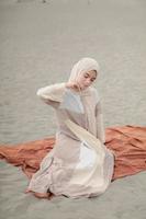 hermosa modelo femenina islámica usando moda hijab, un vestido de novia moderno para mujer musulmana sentada en la arena y la playa. retrata a una modelo asiática usando hiyab divirtiéndose en la playa con árboles foto