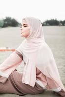 hermosa modelo femenina islámica usando moda hijab, un vestido de novia moderno para mujer musulmana sentada en la arena y la playa. retrata a una modelo asiática usando hiyab divirtiéndose en la playa con árboles foto