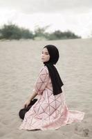 Beautiful islamic female model wearing hijab fashion, a modern wedding dress for muslim woman sitting in the sand and beach. Portrait an asian girl model using hijab having fun at the beach with trees photo