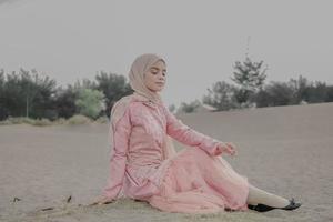 hermosa modelo femenina islámica usando moda hijab, un vestido de novia moderno para mujer musulmana sentada en la arena y la playa. retrata a una modelo asiática usando hiyab divirtiéndose en la playa con árboles foto