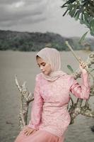 Beautiful islamic female model wearing hijab fashion, a modern wedding dress for muslim woman sitting in the sand and beach. Portrait an asian girl model using hijab having fun at the beach with trees photo