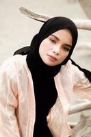 hermosa modelo femenina islámica usando moda hijab, un vestido de novia moderno para mujer musulmana sentada en la arena y la playa. retrata a una modelo asiática usando hiyab divirtiéndose en la playa con árboles foto