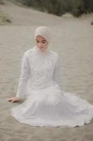 Beautiful islamic female model wearing hijab fashion, a modern wedding dress for muslim woman sitting in the sand and beach. Portrait an asian girl model using hijab having fun at the beach with trees photo