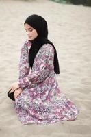 Beautiful islamic female model wearing hijab fashion, a modern wedding dress for muslim woman sitting in the sand and beach. Portrait an asian girl model using hijab having fun at the beach with trees photo