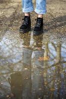 Bottom half of a person's legs wearing boots and standing in front of a puddle of mud in a forest with the person and the trees reflecting in the water.