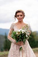 Beautiful bride posing in her wedding dress on a background of mountains. In her hands she hold a bouquet of wildflowers.