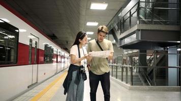 Thai man helps a beautiful Asian female tourist search information and find location for travel at the train station platform, passenger trip lifestyle, casual transportation in journey vacation.