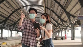 Face mask man helps Asian female tourist search information, find travel locations with tablet at public train station platforms, passenger trip lifestyles, casual journey vacation transportation.
