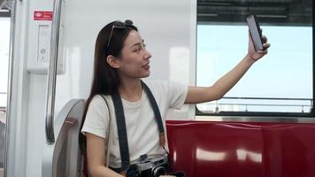 jovem bela turista asiática conversando on-line, chamada de vídeo para celular, check-in e postagem de mídia social, transporte de trem urbano de viagem, estilo de vida de passageiros da cidade, viagem de férias feliz. video
