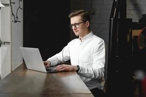 Young man wearing white shirt and eye glasses sitting with laptop and working in cafe photo