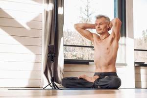 Senior man with a naked torso practices yoga lesson via smartphone in studio. Copy, empty space for text photo