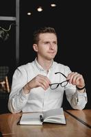 Young man wearing white shirt and eye glasses sitting at the table holding pen takes notes to his agenda on a dark background in office. Copy, empty space for text photo