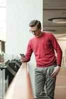 Portrait of a young man with glasses and with a smartphone in his hands near a glass fence in the mall. Communication concept photo