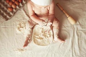 Little boy baker in a chef's hat sitting on the table playing with flour on a brown background with a wooden rolling pin, a round rustic sieve and eggs. Top view. Copy, empty space for text photo