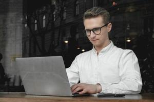 Young man wearing white shirt and eye glasses sitting with laptop and working in cafe. Freelance concept. Copy, empty space for text photo