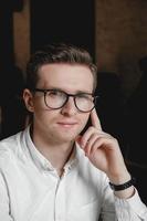 Portrait of a young man in glasses and a white shirt on a dark background. Copy, empty space for text photo