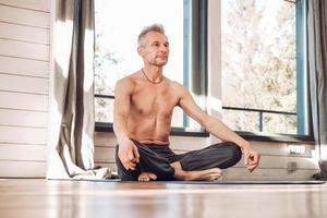 Senior man with a naked torso sitting in a meditative position on a mat in the studio in front of the window. Copy, empty space for text photo