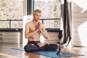 un anciano con un torso desnudo practica una lección de yoga a través de un teléfono inteligente en el estudio. copiar, espacio vacío para texto foto