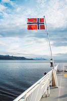 Norway Flag on sea and mountains background. Norwegian flag hanging on the railing of the ship and waving above the water. Norvegian fjord with a flag photo