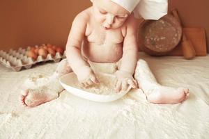 Little boy baker in a chef's hat sitting on the table playing with flour on a brown background with a wooden rolling pin, a round rustic sieve and eggs. Copy, empty space for text photo