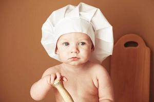 Portrait little baby boy in a chef's hat sitting on the table holds a wooden rolling pin on a brown background. Copy, empty space for text photo