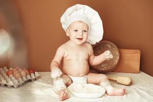 Little boy baker in a chef's hat sitting on the table playing with flour on a brown background with a wooden rolling pin, a round rustic sieve and eggs. Copy, empty space for text photo