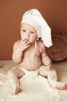 Little boy baker in a chef's hat sitting on the table playing with flour on a brown background with a wooden rolling pin, a round rustic sieve and eggs. Copy, empty space for text photo