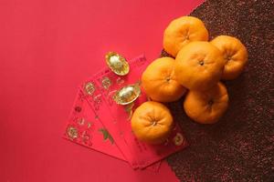 Chinese New Year festival concept. Mandarin oranges, red envelopes, and gold ingots on red cloth with red background. photo
