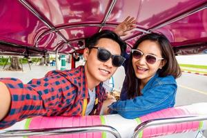 Cheerful young Asian couple tourists taking selfie while traveling on local colorful Tuk Tuk taxi exploring Bangkok city, Thailand photo
