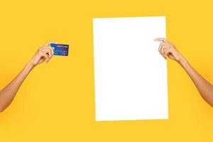 Female hands holding empty white billboard paper and credit card on isolated studio yellow bakground photo