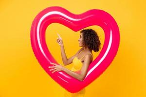 retrato de vista lateral de una mujer afroamericana sonriente apuntando hacia arriba sosteniendo un globo con forma de corazón rosa aislado en un fondo amarillo foto