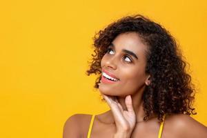 primer plano retrato de una hermosa mujer afroamericana sonriente pensando en mirar hacia arriba aislada en un fondo amarillo foto