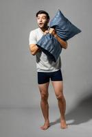 Full lenght portrait of young Caucasian man holding pillow and being shocked after waking up, studio shot in light gray isolated background photo