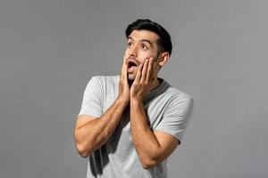 Shocked young Caucasian man with hands on cheeks gasping and looking up in light gray isolated studio background photo