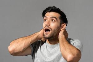 Stunned young Caucasian man gasping with hands around neck looking upward on light gray studio background photo