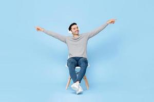 retrato de un relajante joven apuesto hombre caucásico sentado en una silla con auriculares escuchando música con los brazos levantados en un estudio aislado de fondo azul foto