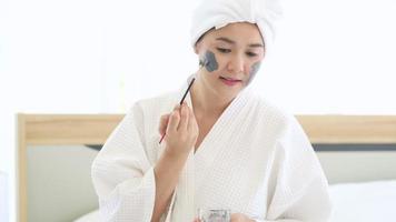 Happy beautiful woman in white bathrobe applying a face mask, skin care and treatment concept video