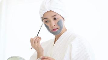 Happy beautiful woman in white bathrobe applying a face mask, skin care and treatment concept video